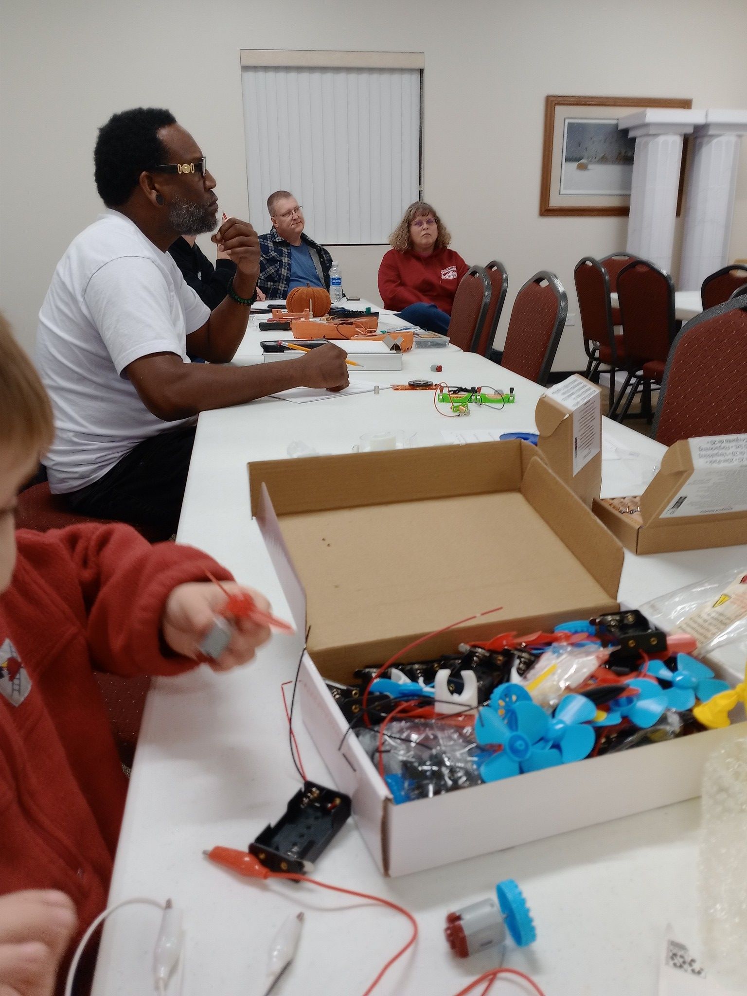 People at a table working with electrical components, in a workshop setting. A child in red jacket plays with a component.