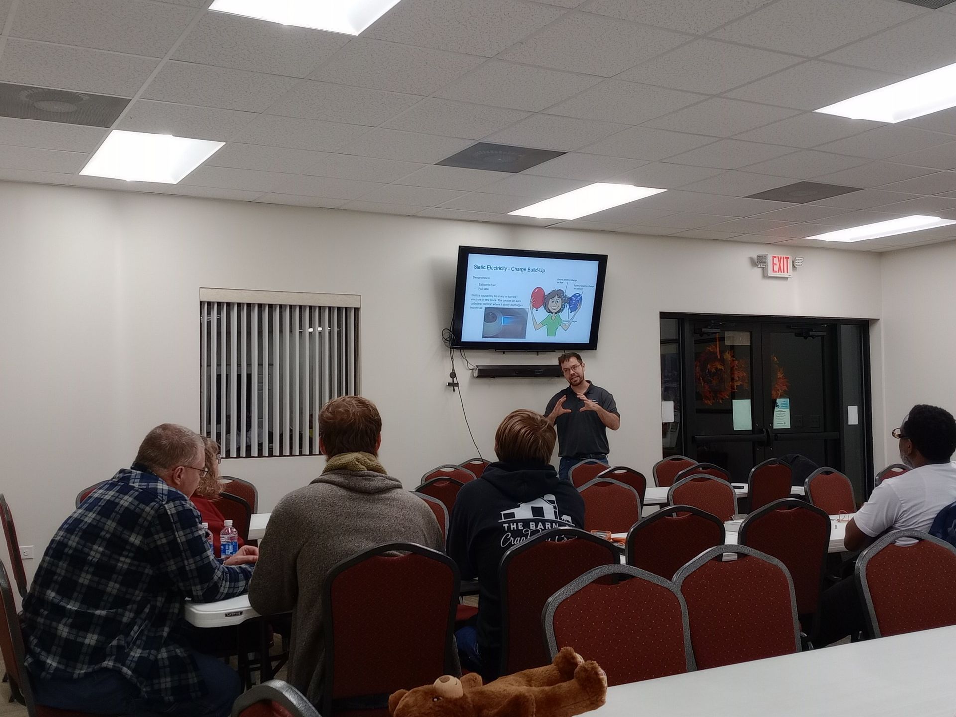 Man presents to a group in a brightly lit room with a TV screen displaying a presentation. Attendees sit in chairs.