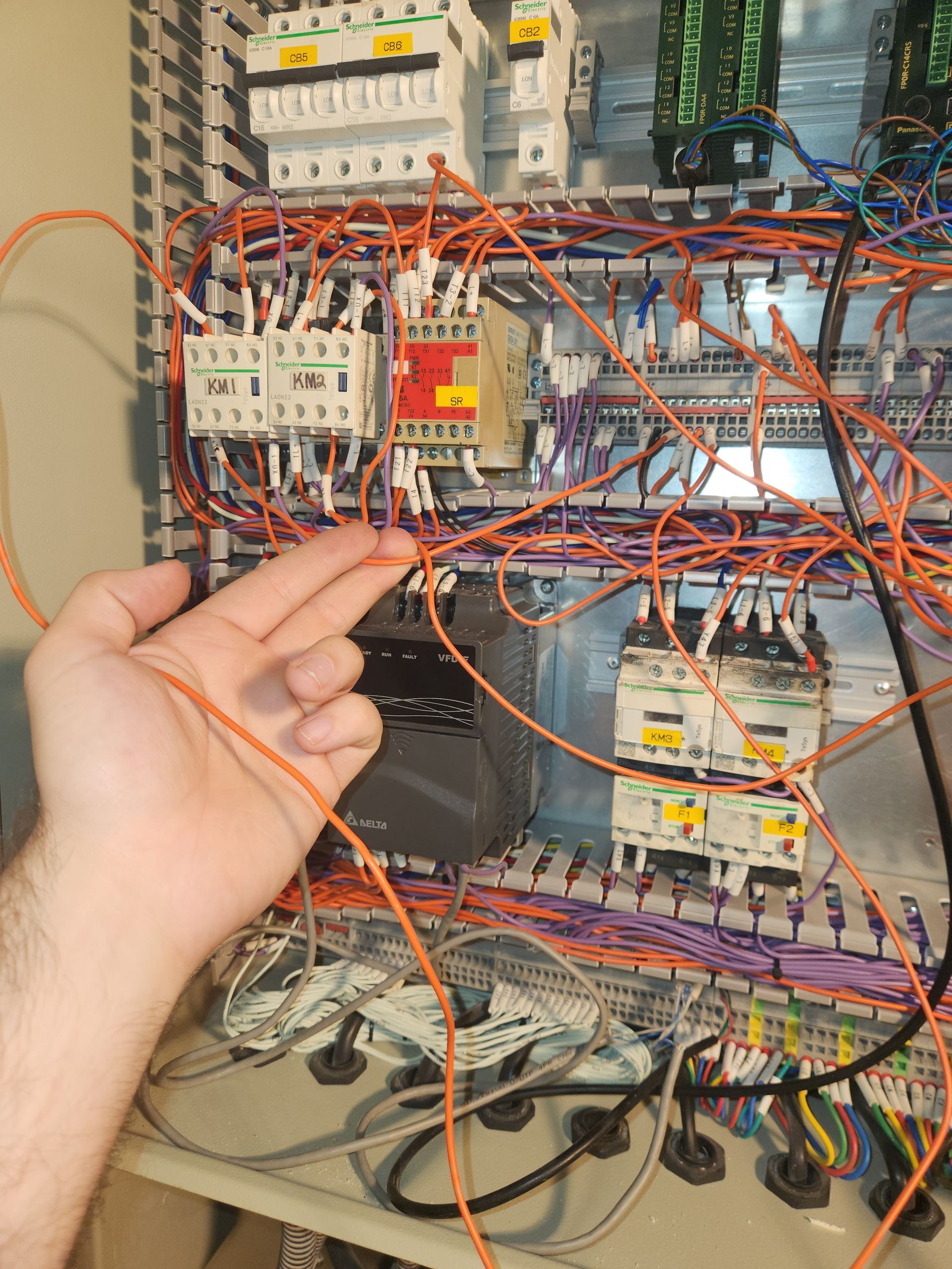 Hand pointing at tangled orange wires within a gray electrical panel.