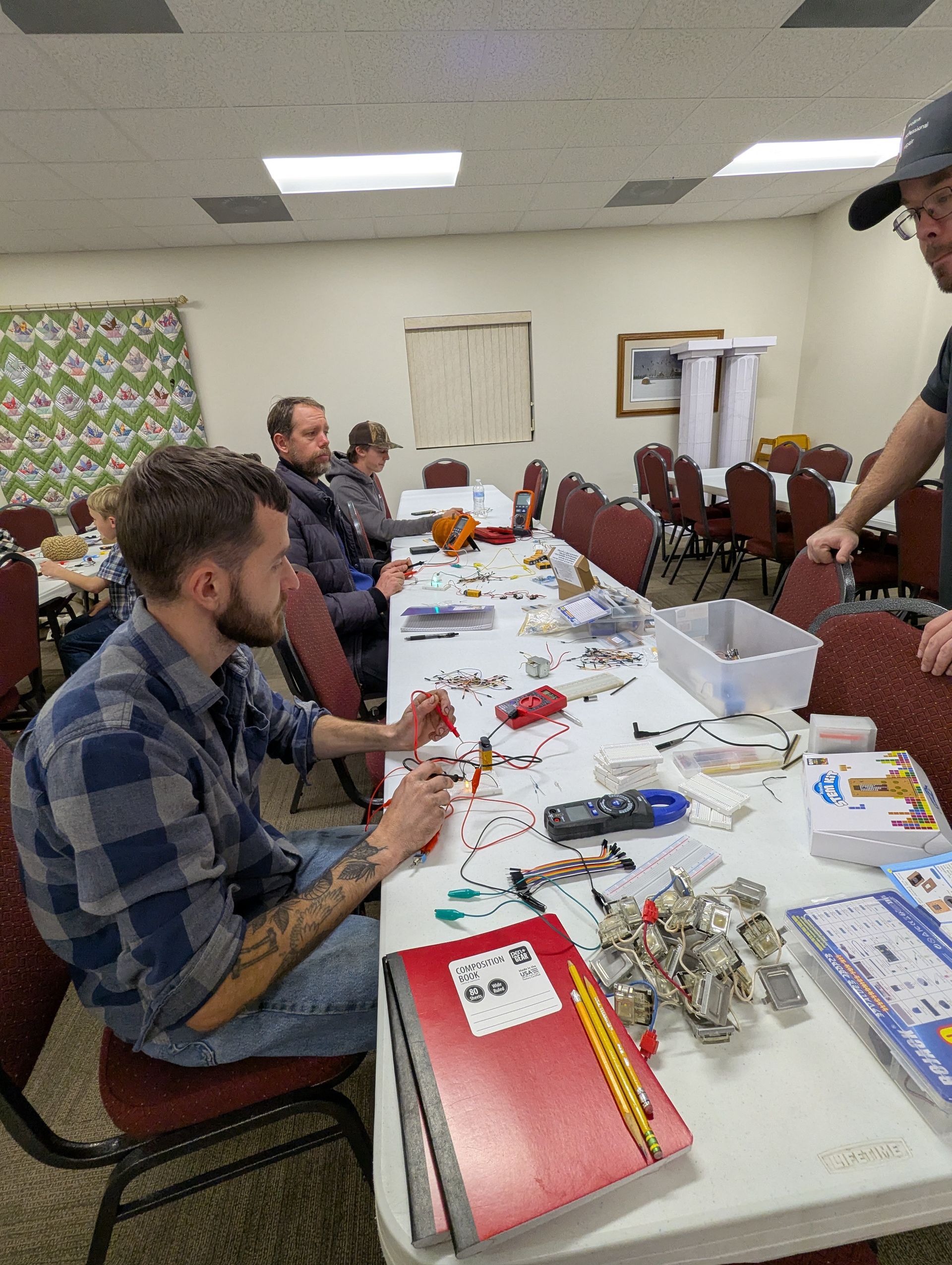 People building with LEGOs at a table. One man in a plaid shirt. Others seated. Supplies scattered.