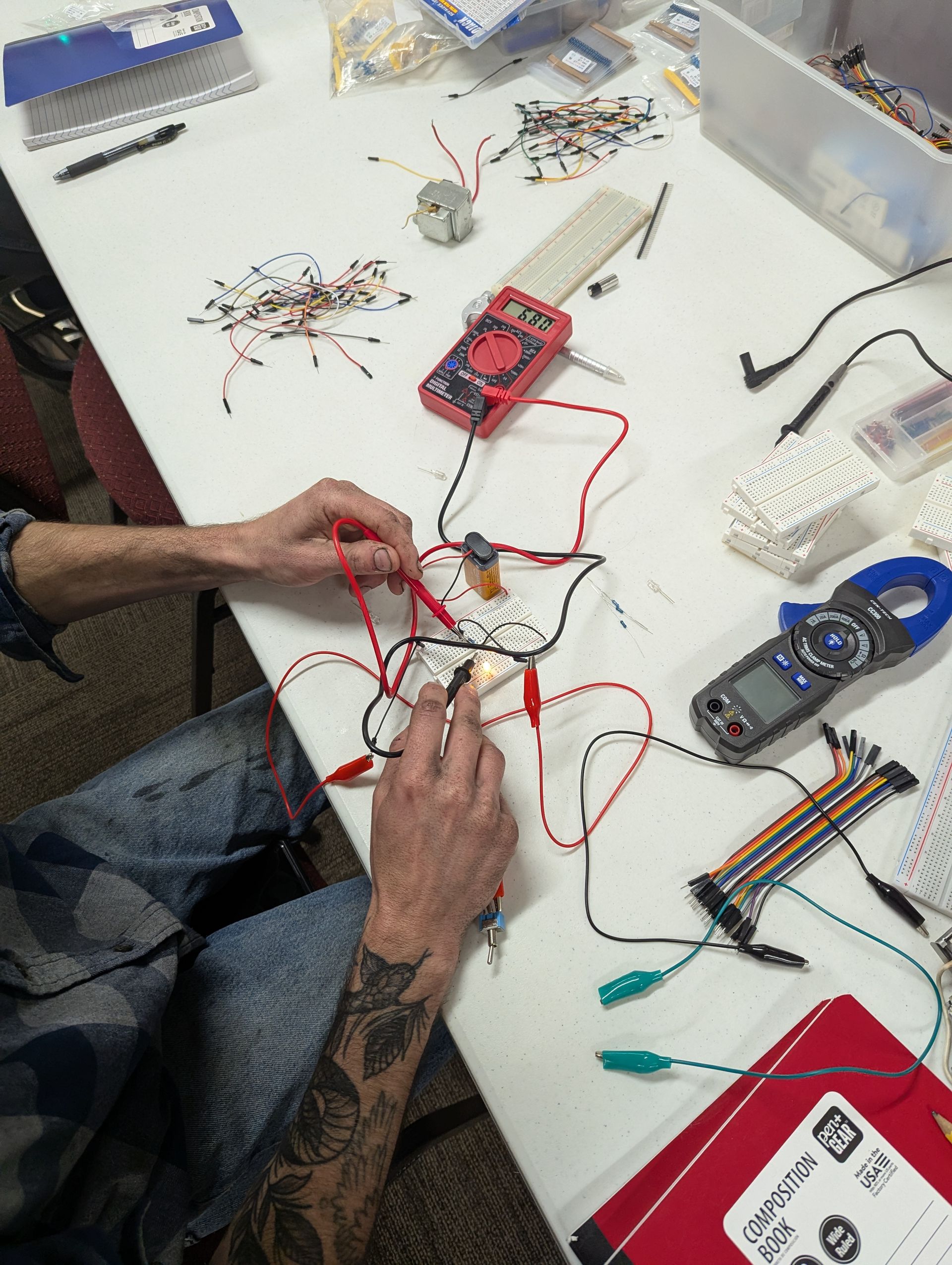 Person working on an electrical circuit with wires, a multimeter, and other components on a white table.