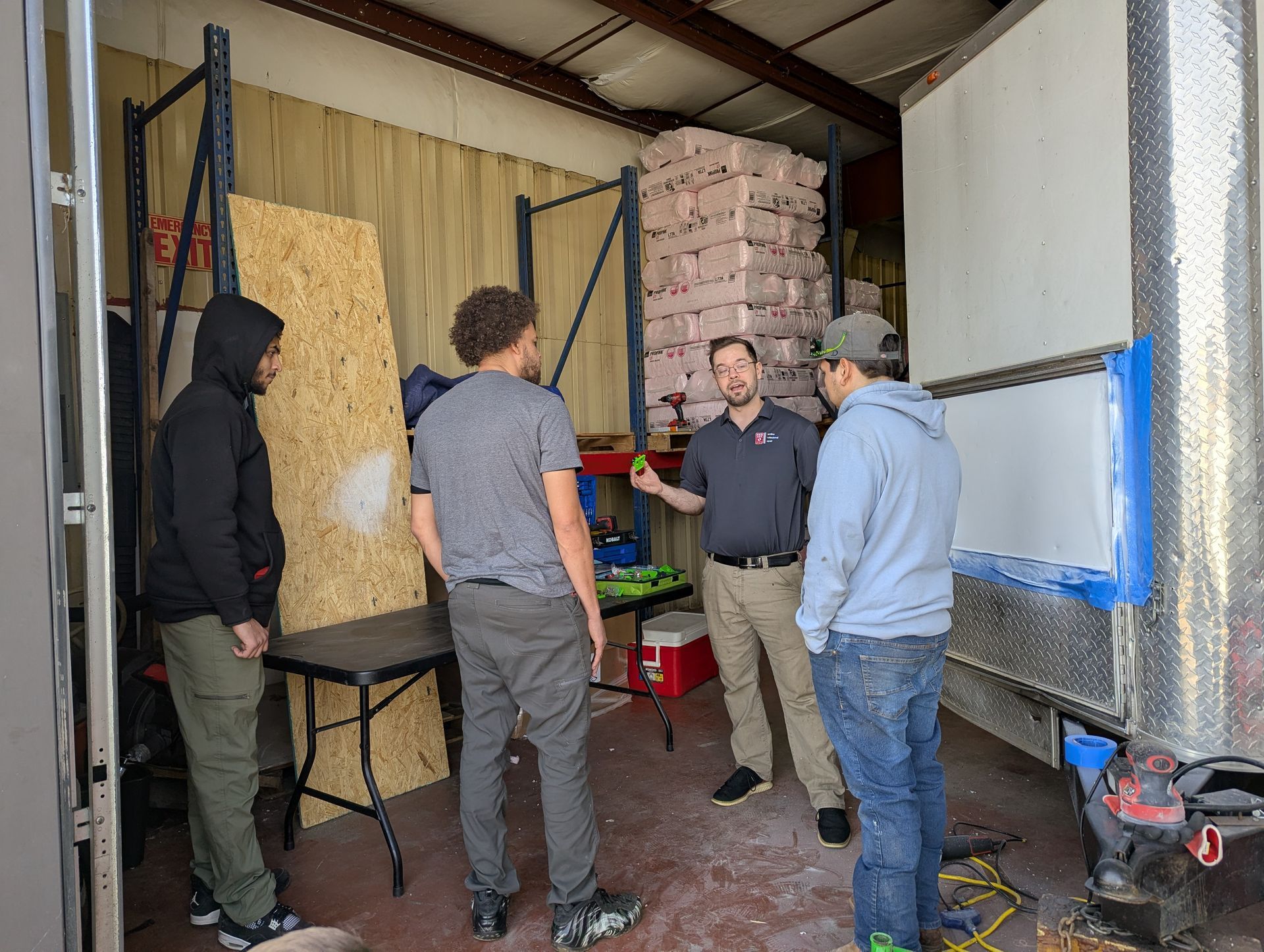 Four men inside a truck, one gesturing. A whiteboard and wood panel are present.