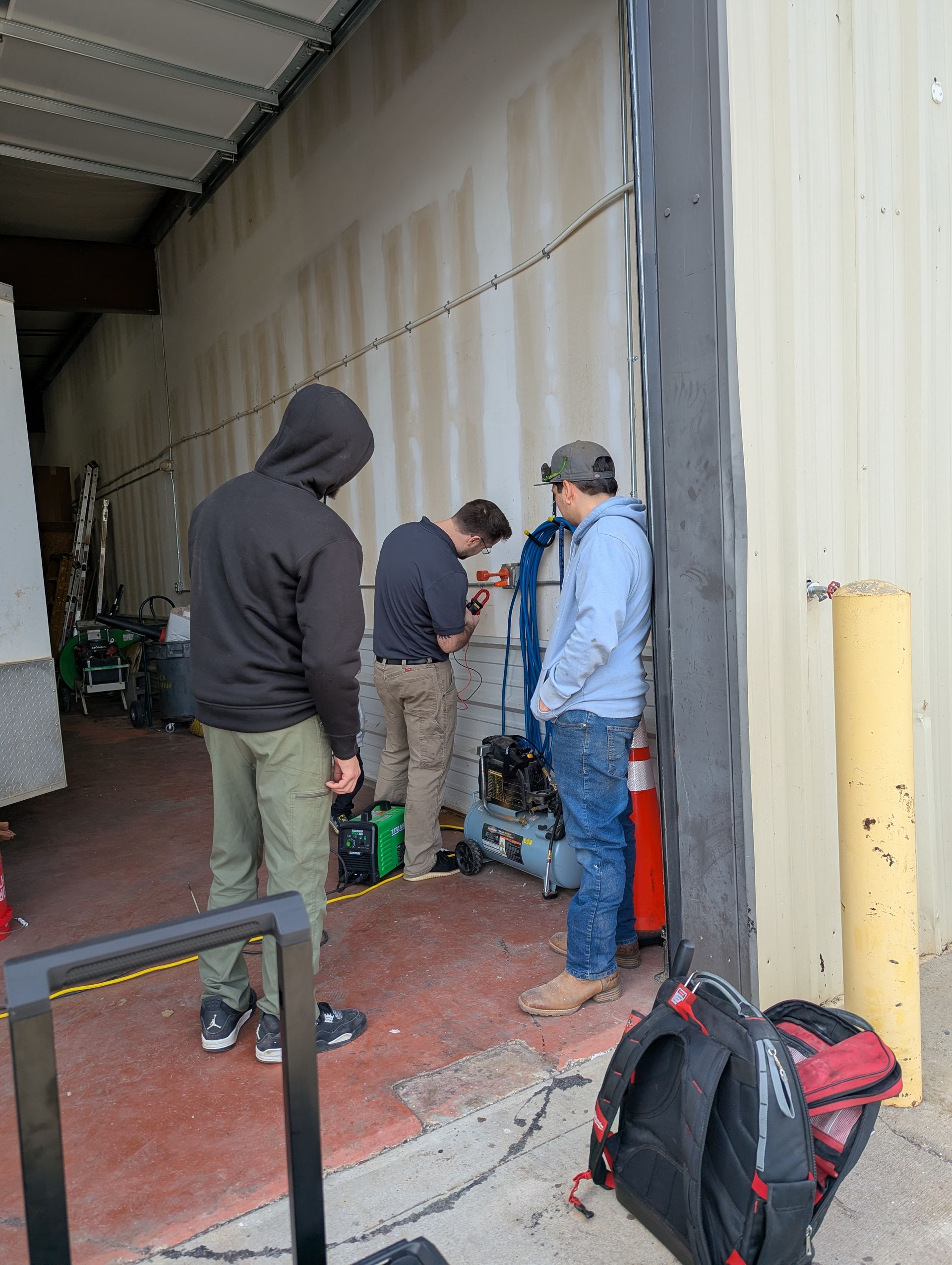 Three people working on equipment by a garage door. One adjusts a valve, the others observe.