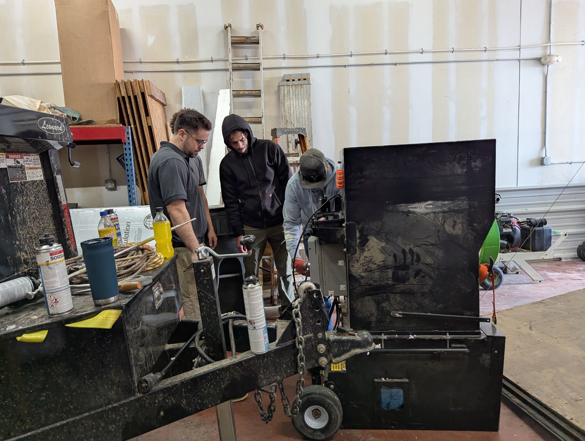Three people inspecting a machine in a workshop. One man works on the machine while the others observe.