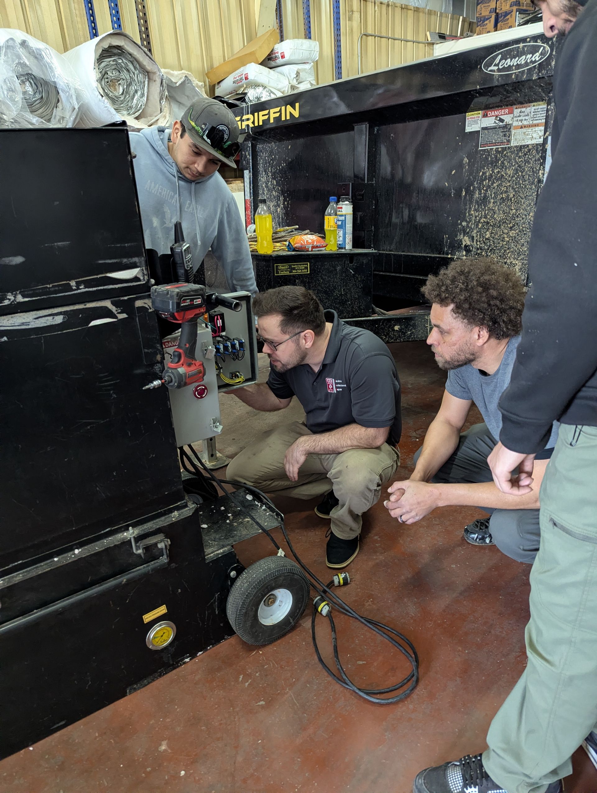 Three men examine machinery inside a workshop. One man points; another peers at the machine.