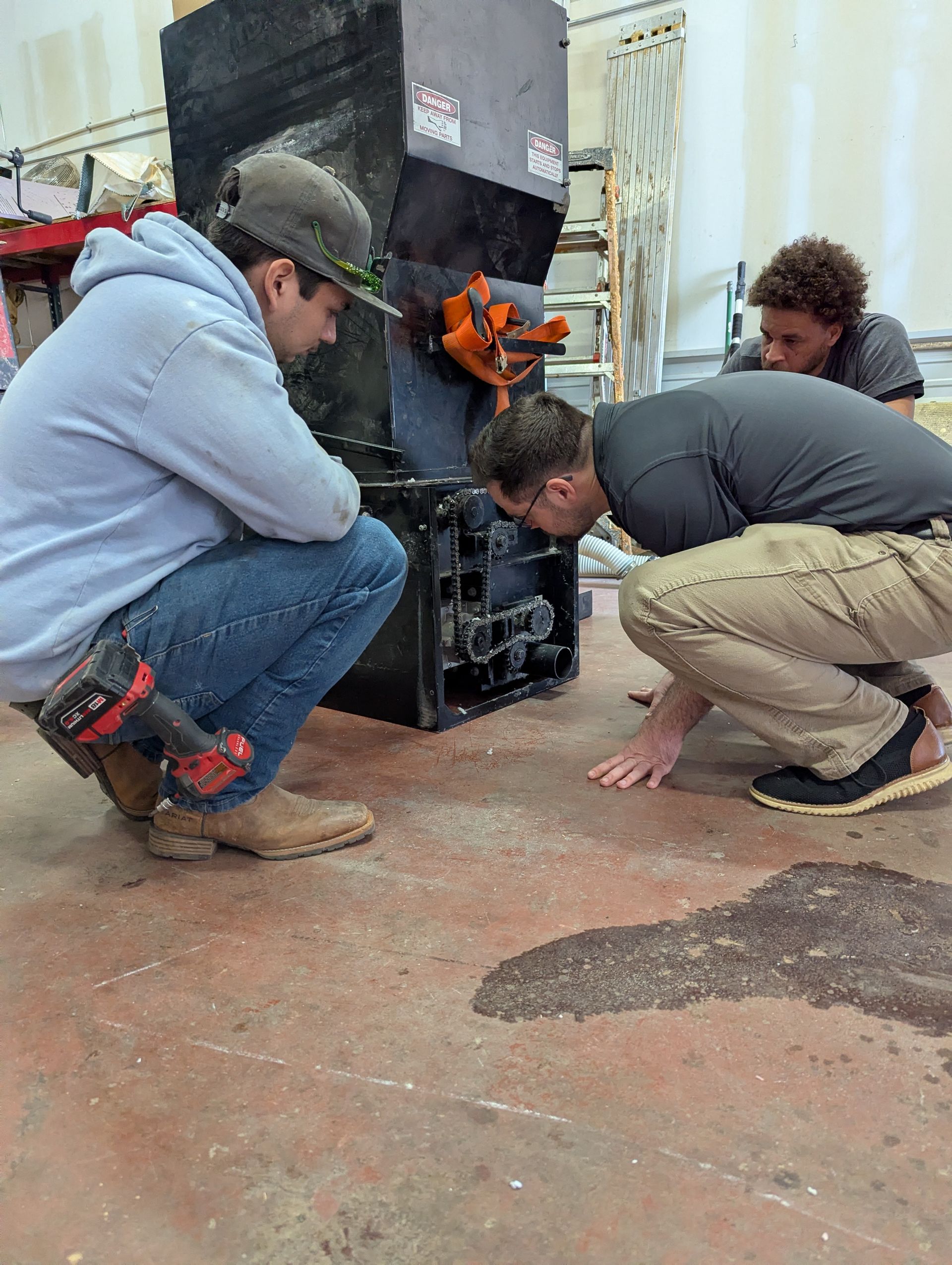 Three people examining machinery on a workshop floor. Two are crouching, one has a drill.