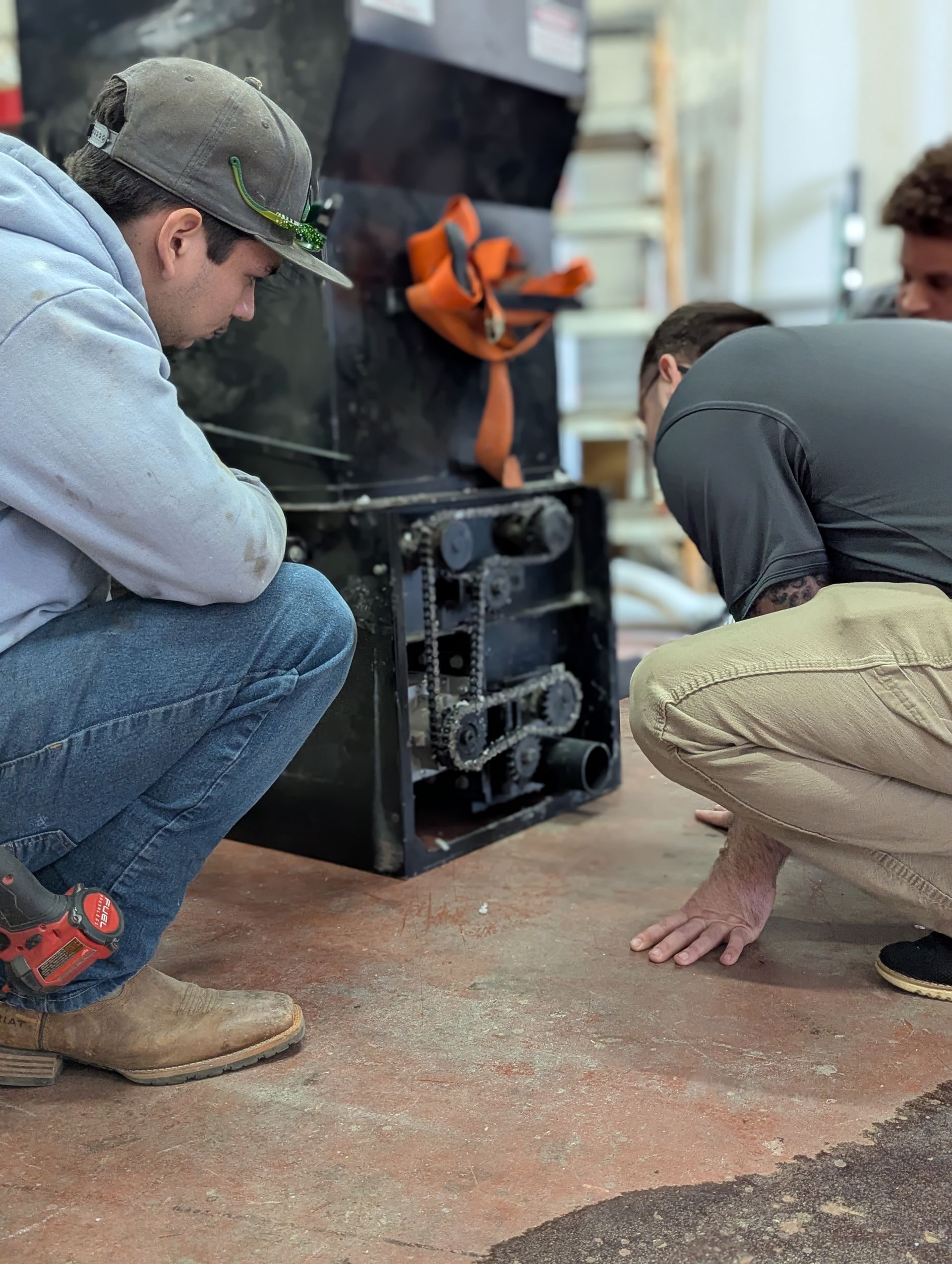 Three people examine the underside of a black machine on a concrete floor.