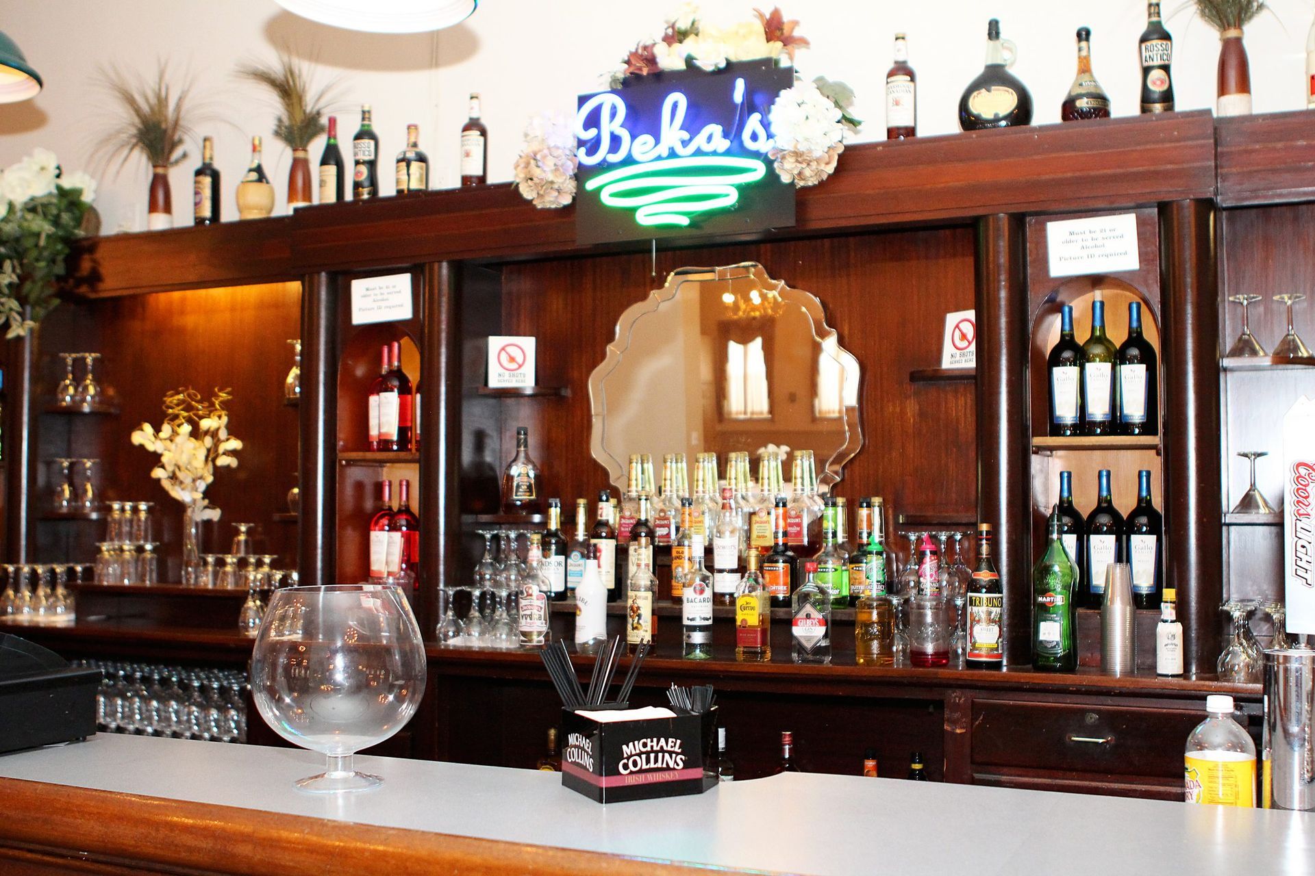 Bar interior with bottles on shelves, drinks on the counter, and a neon sign that reads