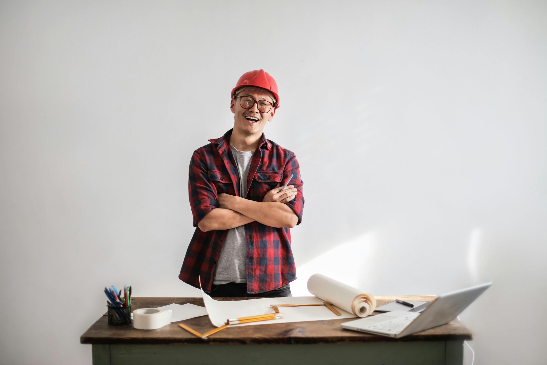 A man wearing a hard hat is standing in front of a desk with his arms crossed.