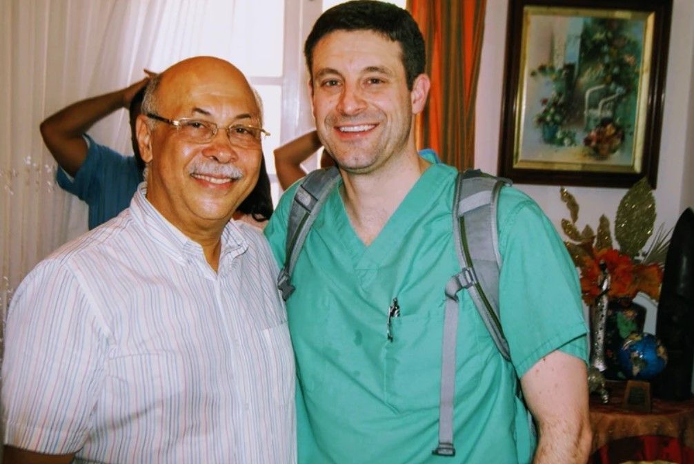 A man in a green scrub top stands next to another man in a white shirt