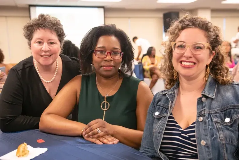 Three women are posing for a picture while sitting at a table.