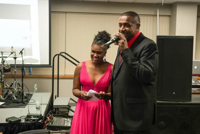 A man is speaking into a microphone next to a woman in a pink dress.
