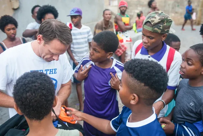A man in a white shirt with the word the on it is talking to a group of young boys.