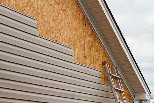 Tan vinyl siding is being installed on the side of a house, leaving exposed wooden sheathing near the roofline and a ladder.