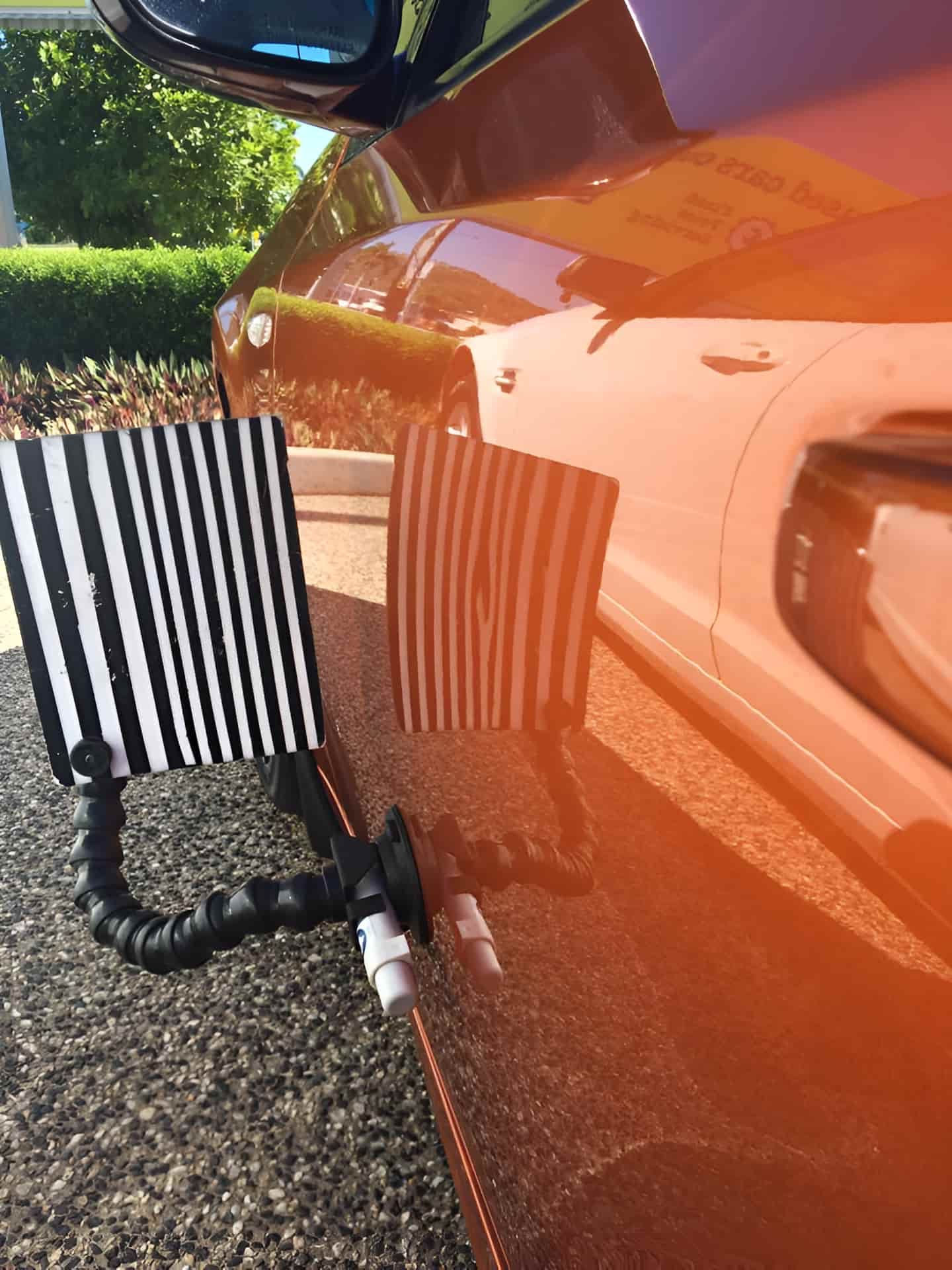 A Black and White Striped Board is Attached to the Side of an Orange Car — Dek-A-Dent Townsville In Wulguru, QLD