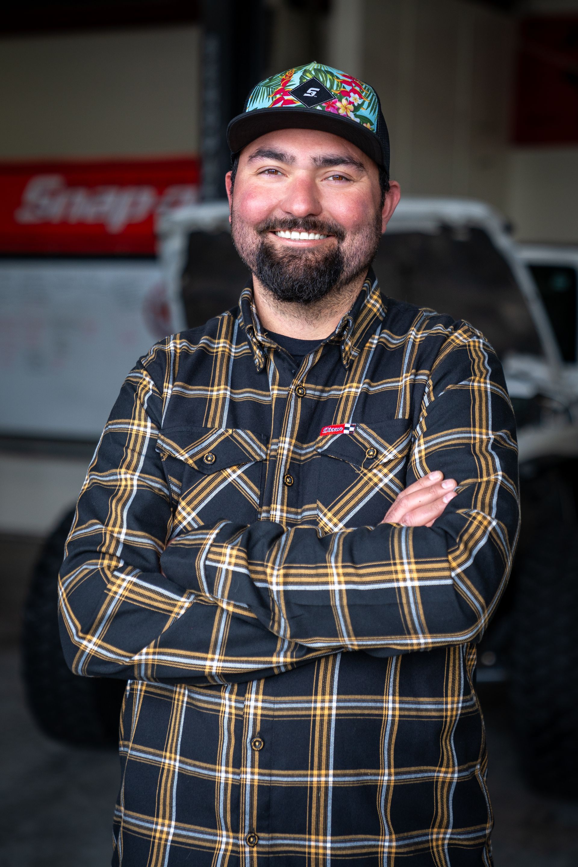 A man in a plaid shirt and hat is standing with his arms crossed.