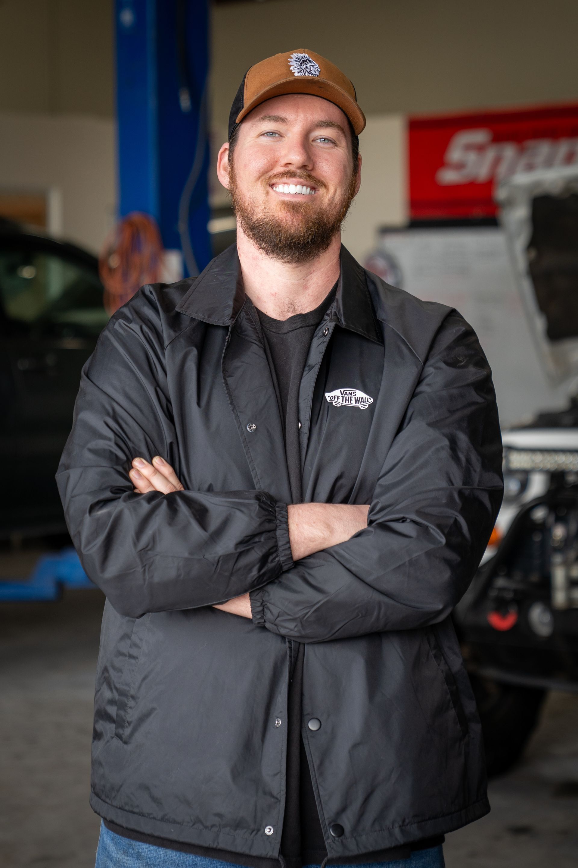A man wearing a hat and a black jacket is standing with his arms crossed in a garage.
