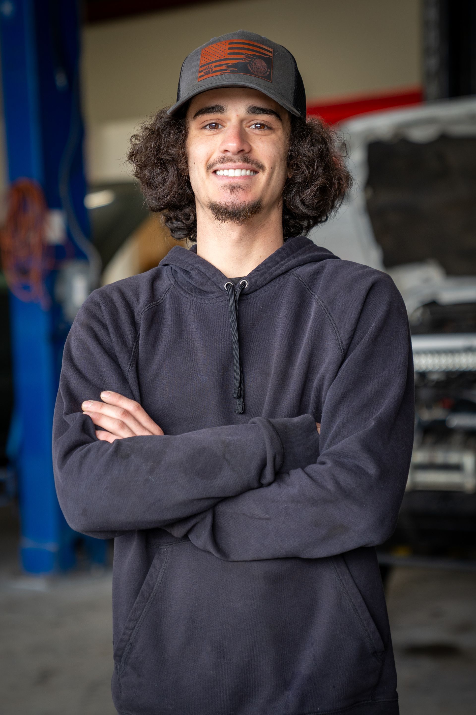 A young man wearing a hat and a hoodie is standing in a garage with his arms crossed.