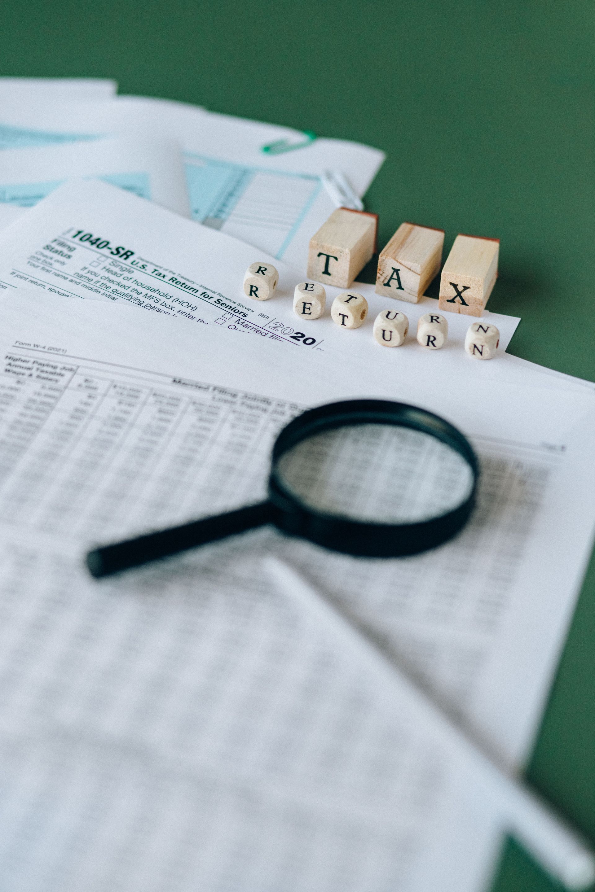 Magnifying glass over tax forms with letter blocks spelling “TAX” on a green desk