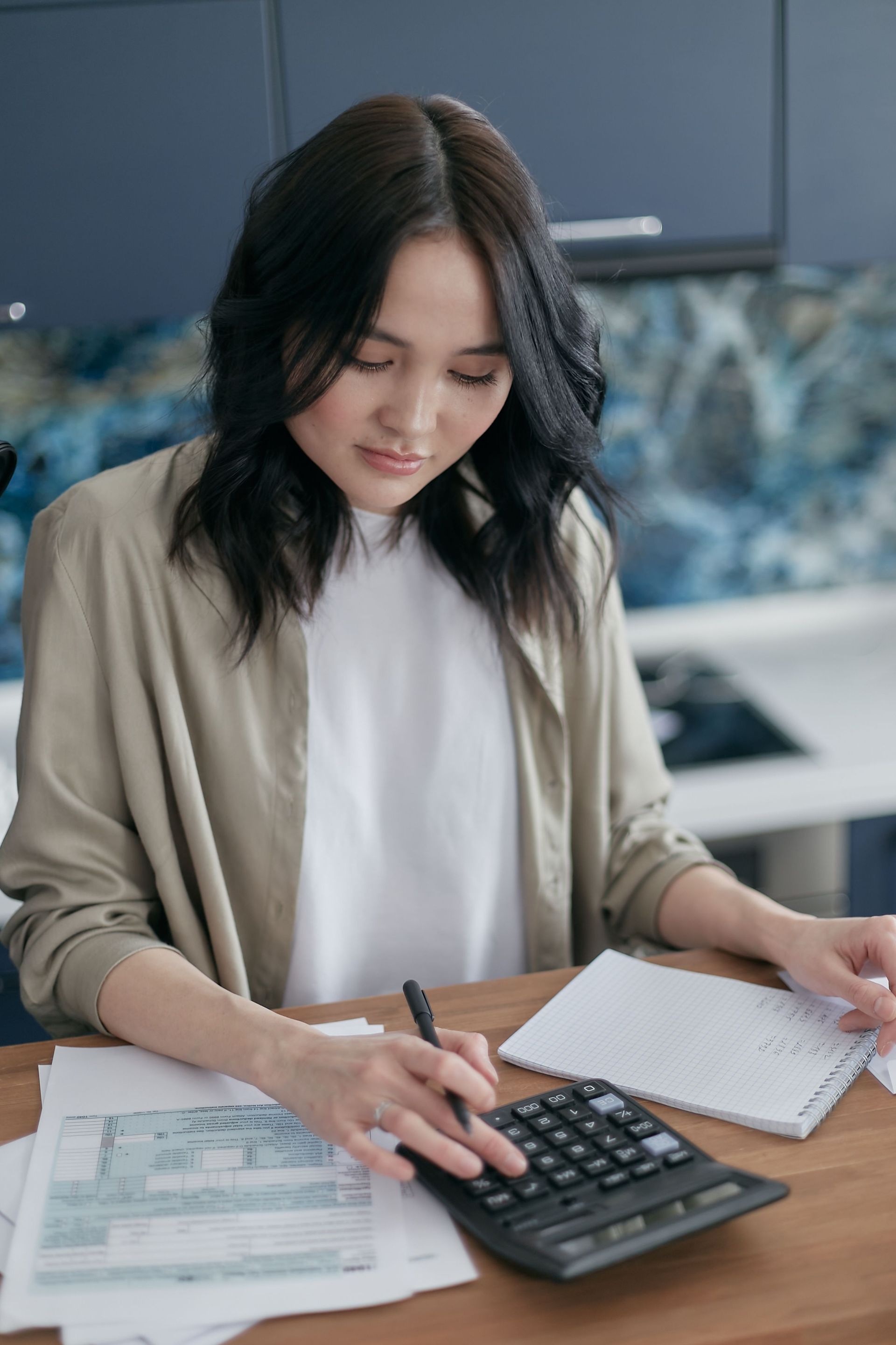 Person at a table using a calculator and reviewing bills with a notebook nearby.