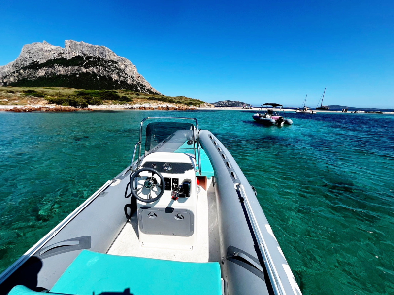 Prua di una barca su acque turchesi vicino a un'isola rocciosa sotto un cielo azzurro brillante.