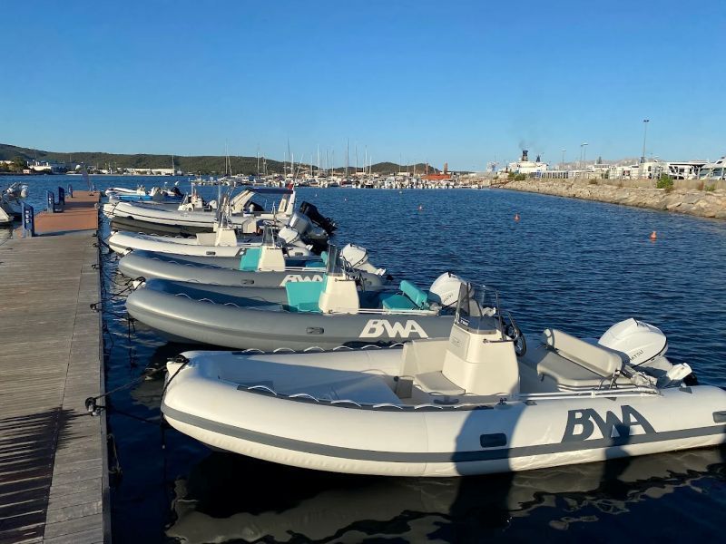 Una fila di gommoni bianchi ormeggiati lungo un lungomare soleggiato con acqua azzurra e la costa in lontananza.