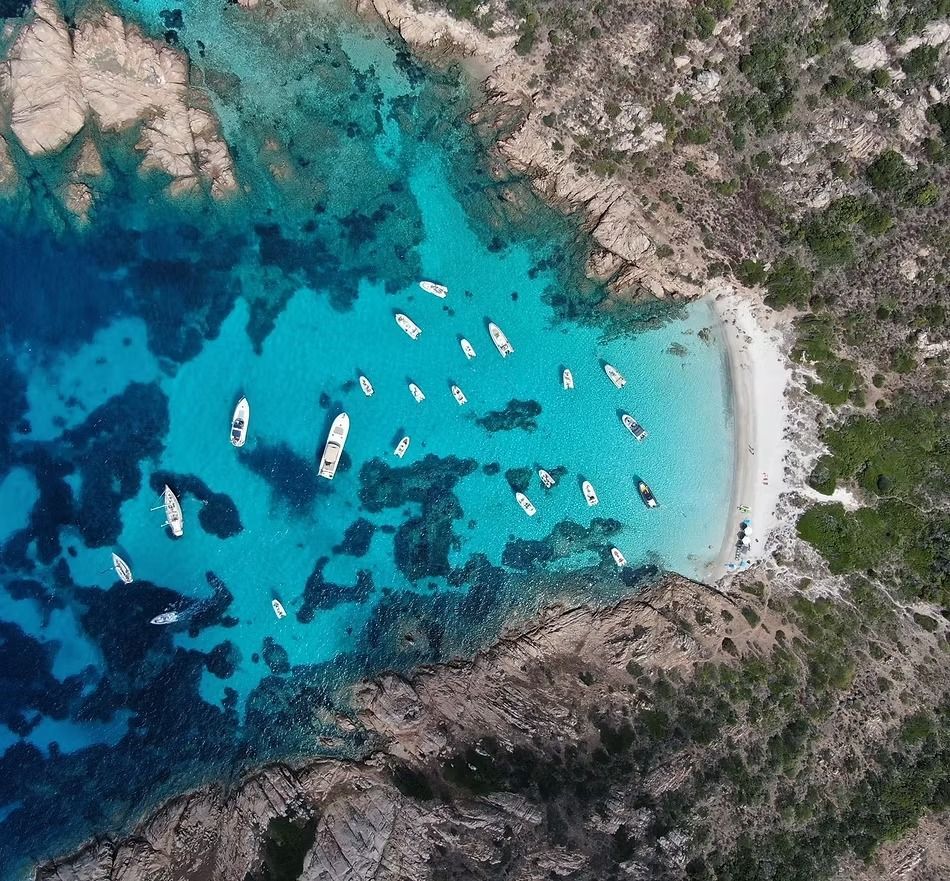 Veduta aerea di una baia turchese con barche ancorate accanto a una piccola spiaggia di sabbia bianca e a una costa rocciosa.