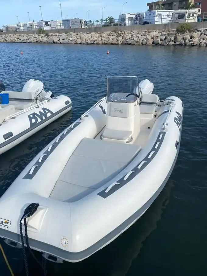 Un motoscafo gonfiabile bianco è ormeggiato in un porto, con un'altra imbarcazione accanto e una diga frangiflutti rocciosa sullo sfondo.