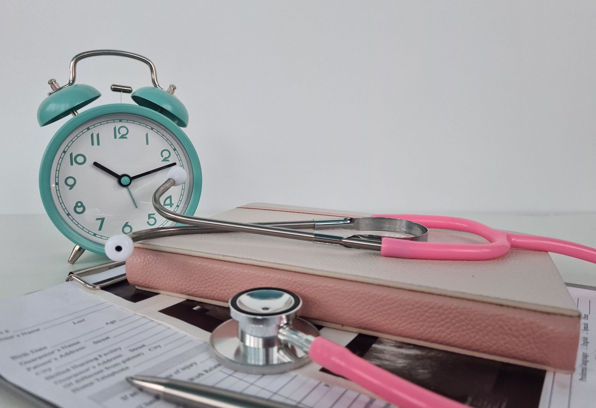 A pink stethoscope is sitting on top of a notebook next to an alarm clock.