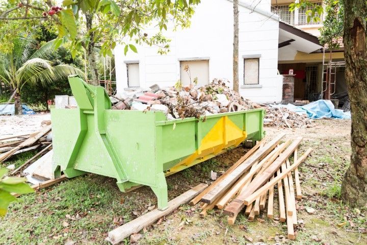 A green dumpster is sitting next to a pile of wood in front of a house.