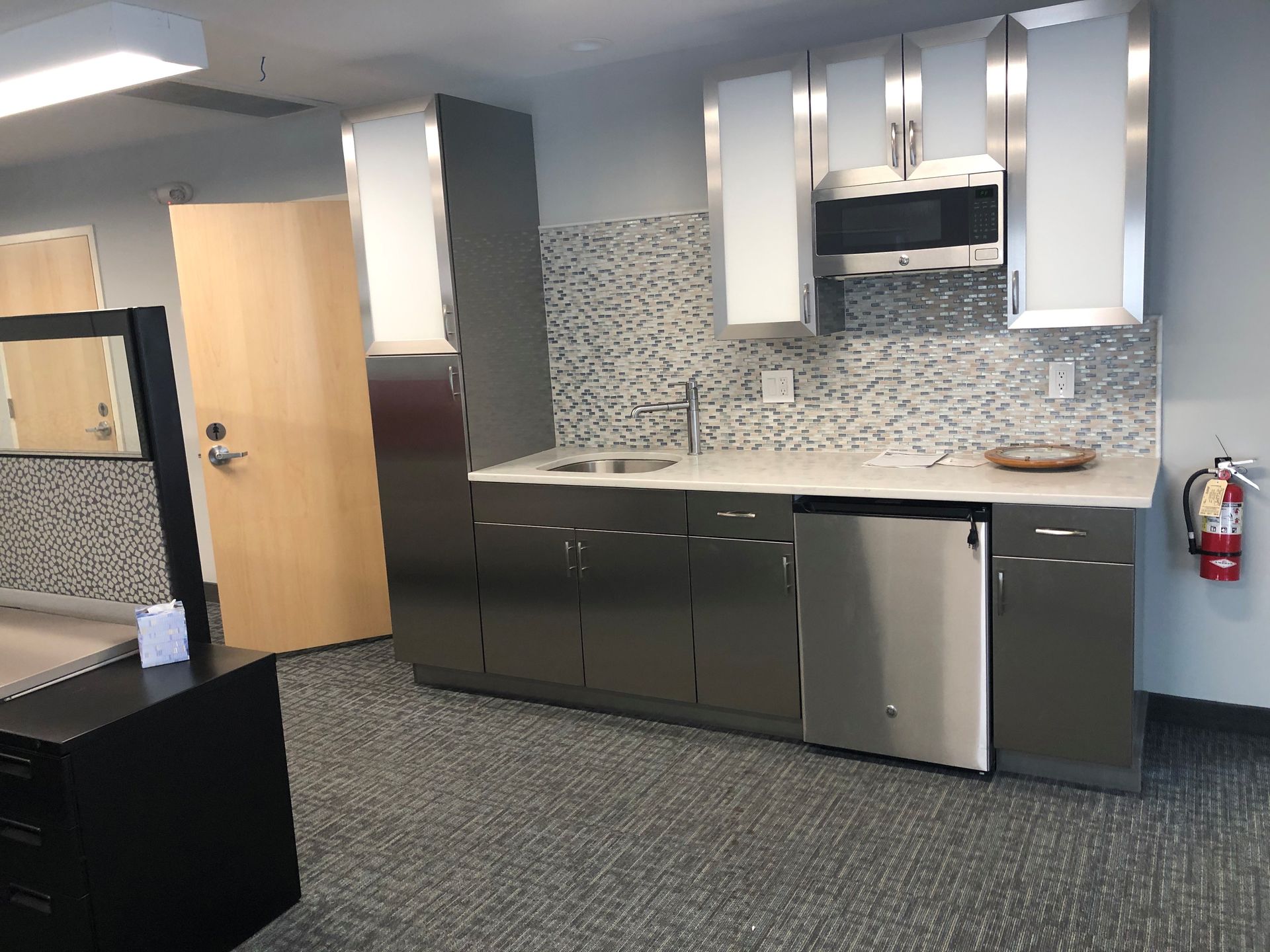 Small office kitchenette with stainless steel cabinets and appliances, sink, microwave, and backsplash.