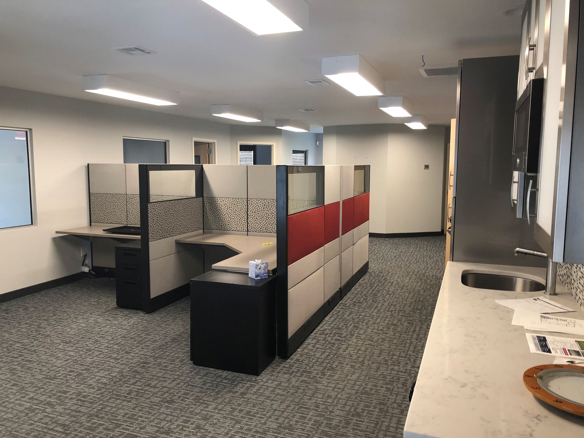 Office space with cubicles, desks, and a kitchenette area. Gray carpet, white walls, and bright overhead lights.