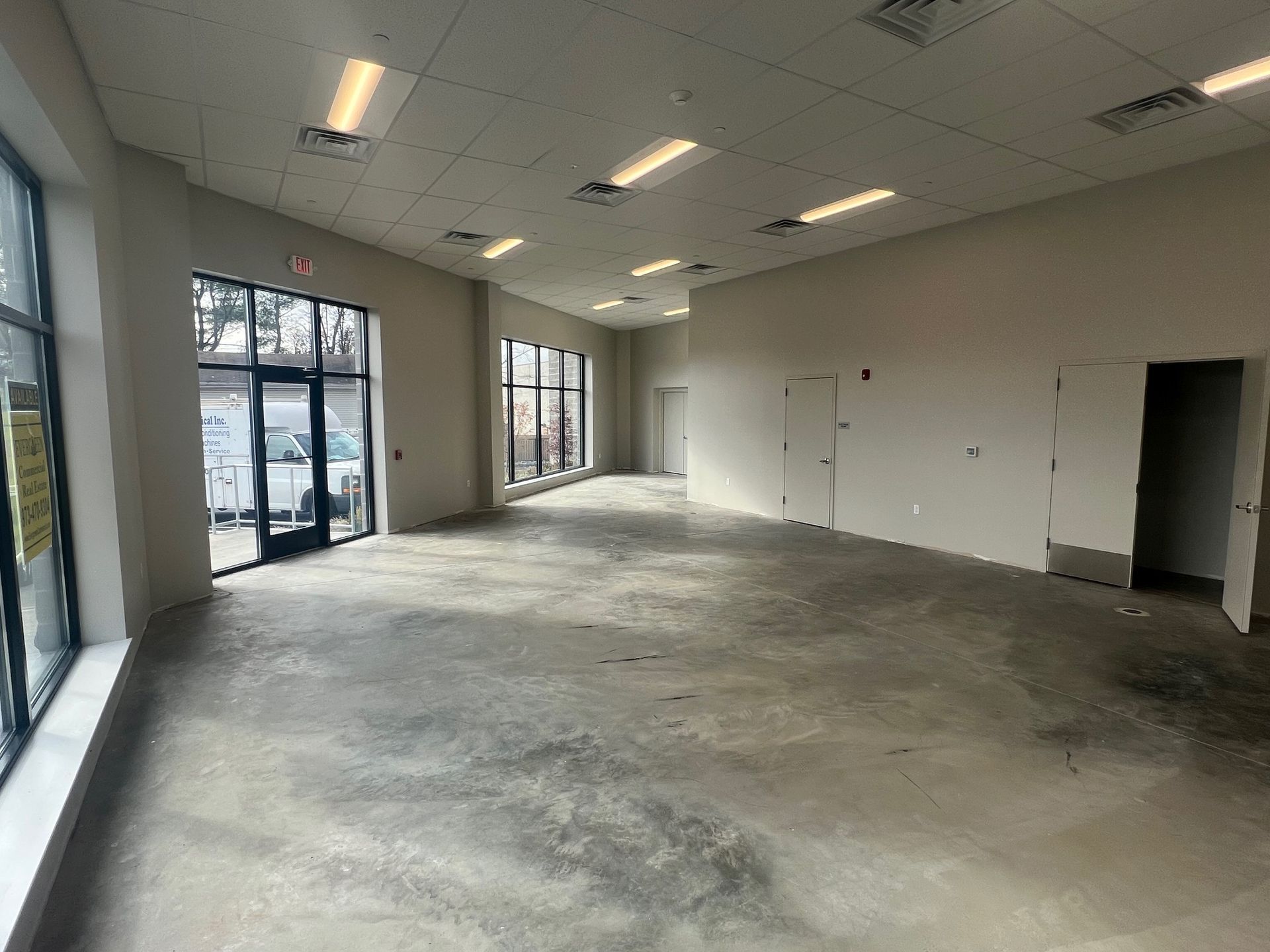 Empty commercial space with large windows and doors, concrete floor, and white walls.