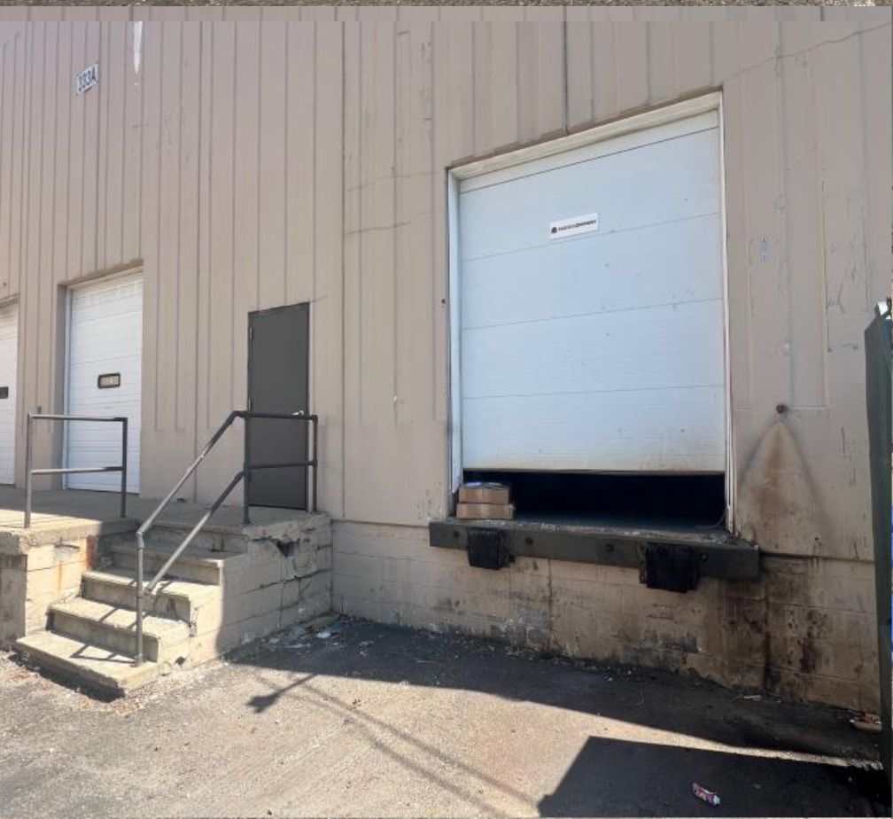 Exterior view of a building with a loading dock, open white door, and steps with a railing.