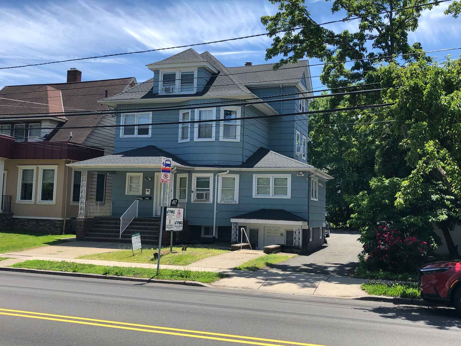 Blue two-story house with white trim and porch beside a road, with trees and a parked red car.