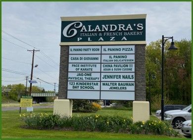 A directory sign for Calandra’s Plaza, listing various businesses, set against a backdrop of trees and a cloudy sky.