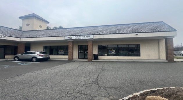 A tan, single-story commercial strip mall building with a grey shingled roof, a silver SUV parked in front, and a sign.