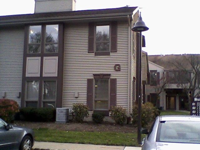 Tan, two-story apartment building with brown shutters and trim, a dark lamppost, and parts of parked cars in front.