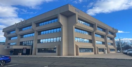 Four-story beige office building with many windows under a blue sky, parked cars in front.