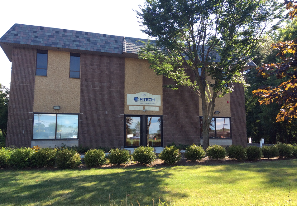 Four-story beige office building with many windows under a blue sky, parked cars in front.