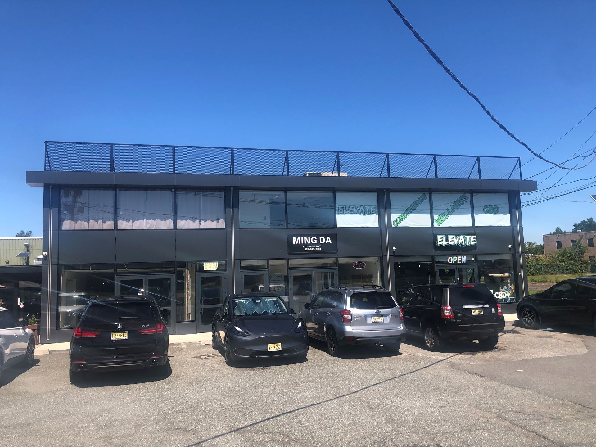 Black building with multiple storefronts, cars parked out front, sunny day.