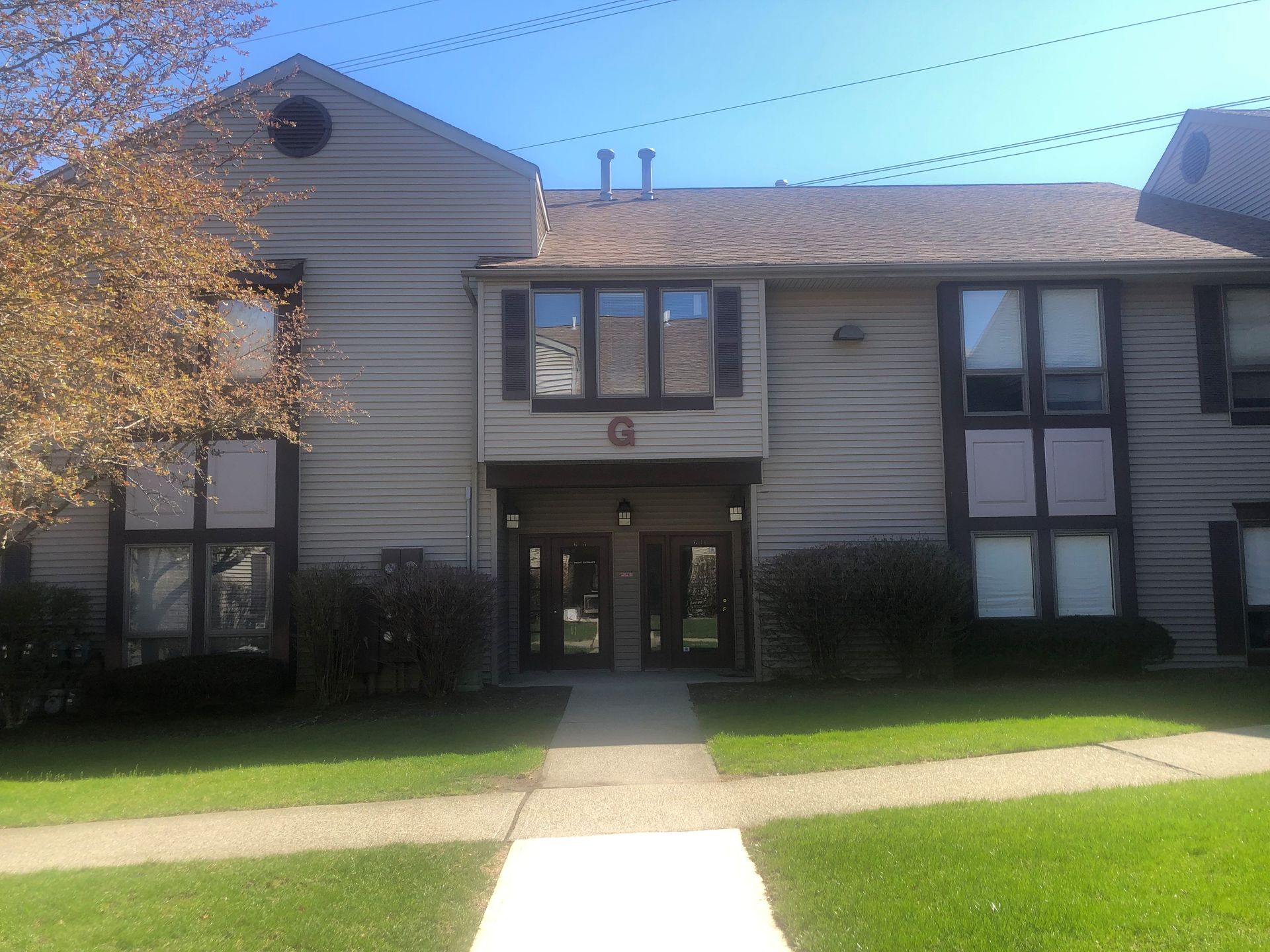 A two-story apartment building with beige siding, dark-trimmed windows, a central entrance labeled C, and a front lawn.