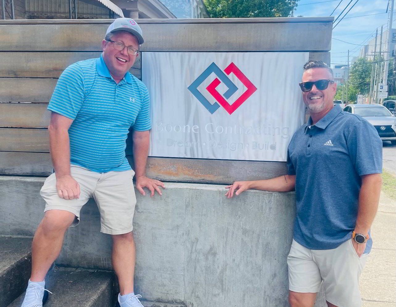Two men are standing next to each other in front of a sign for Boone Contracting