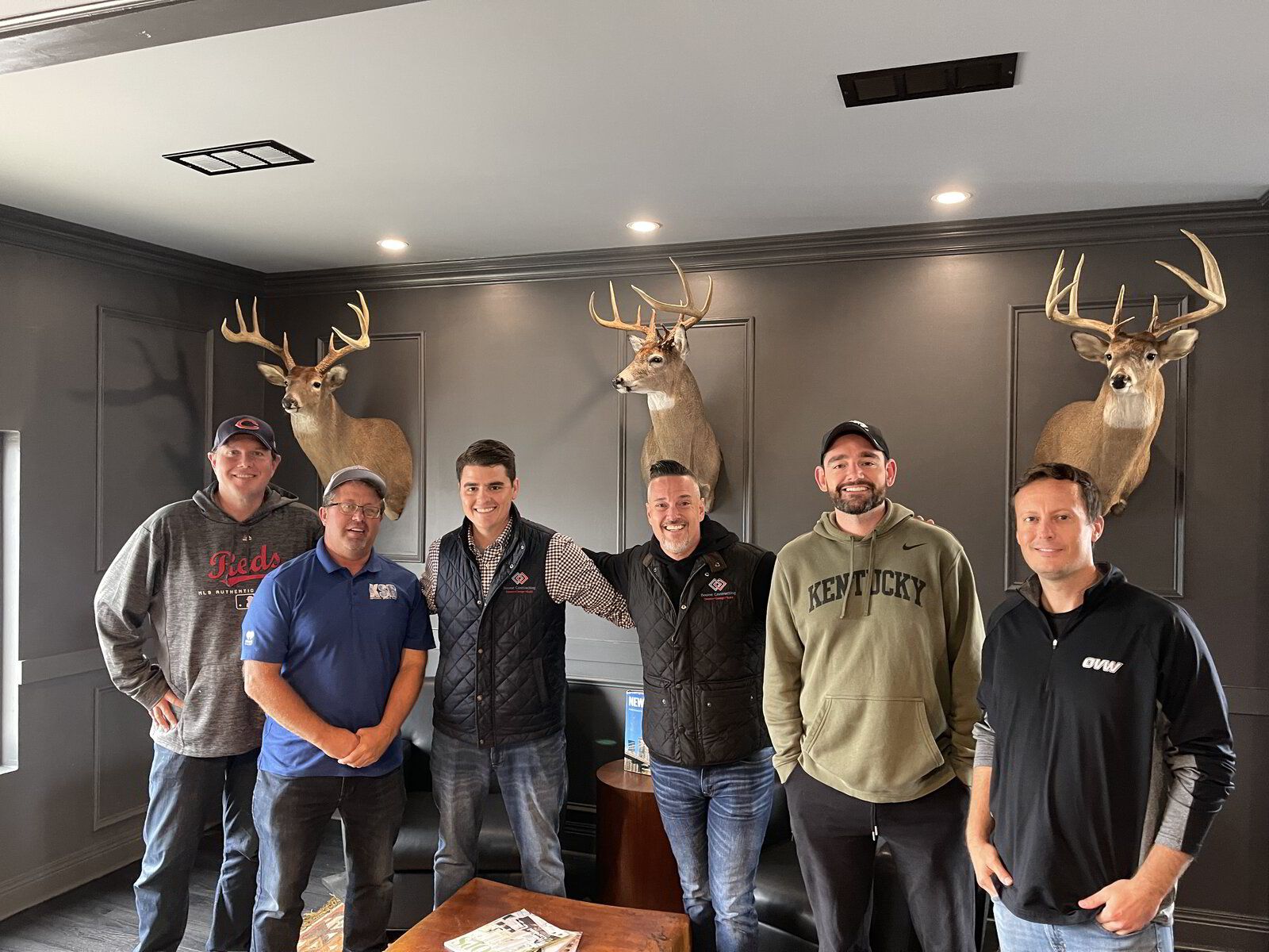 A group of men are posing for a picture in a living room with deer heads on the wall.