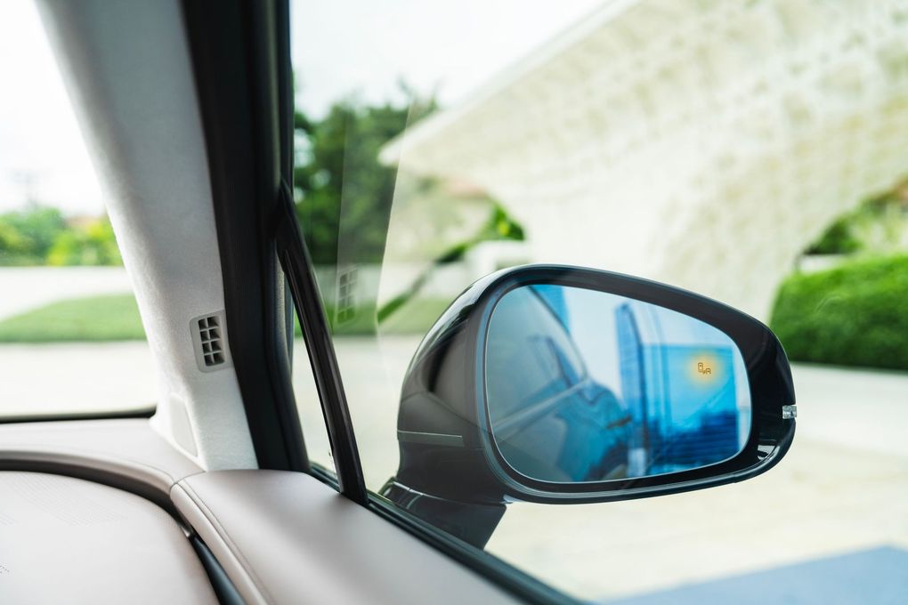 Car side mirror with a blind spot indicator, reflecting a blue sky and white building.