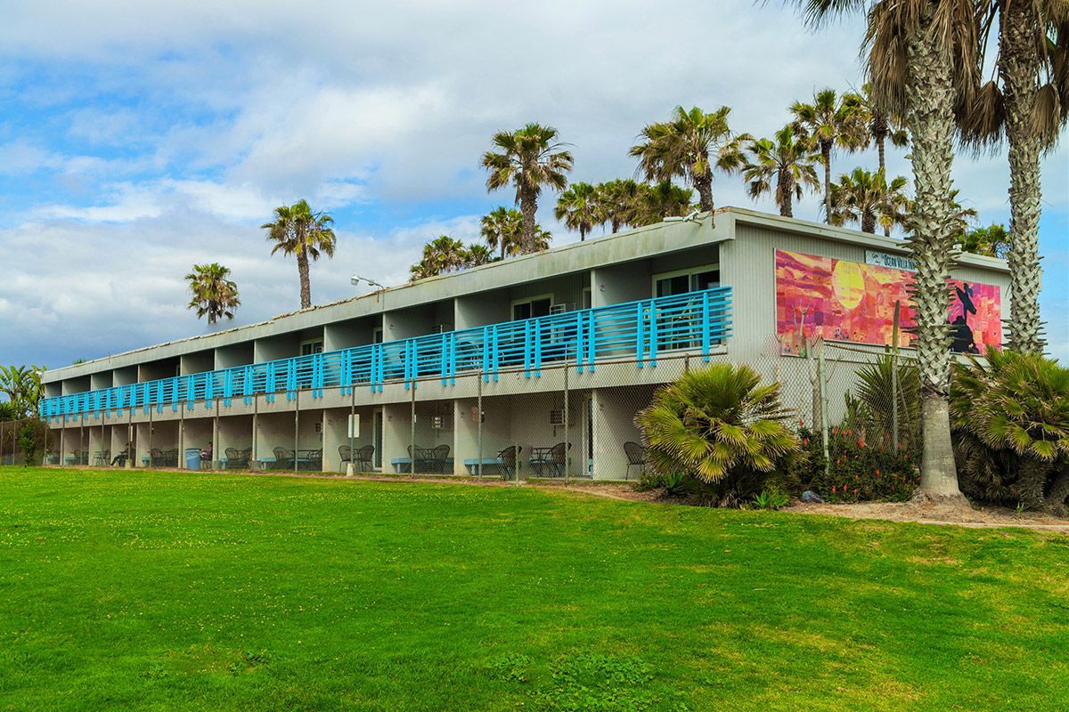 Motel with teal balconies, mural, palm trees, green lawn, cloudy sky.