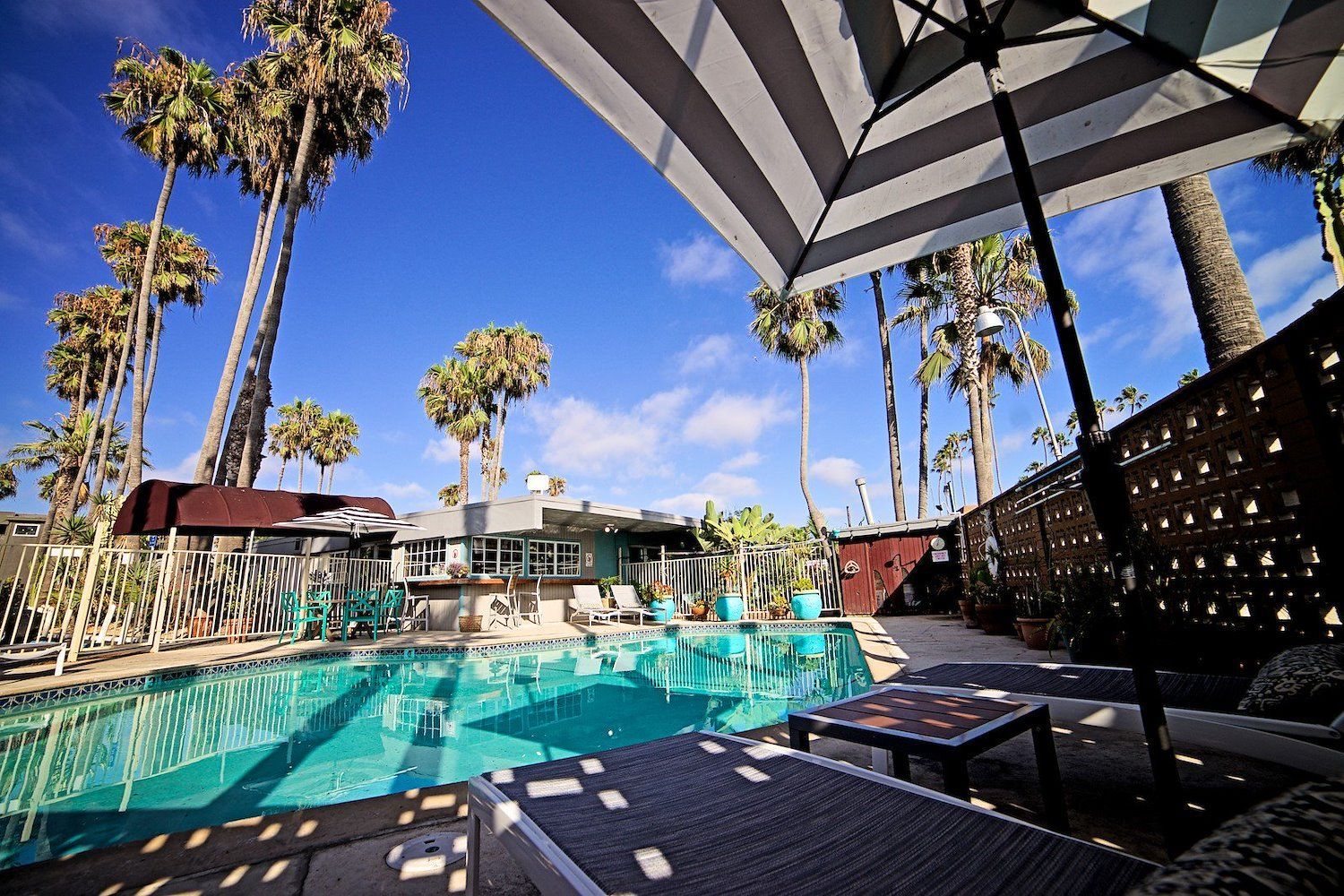 Poolside scene with a turquoise pool, palm trees, and a striped umbrella against a blue sky.