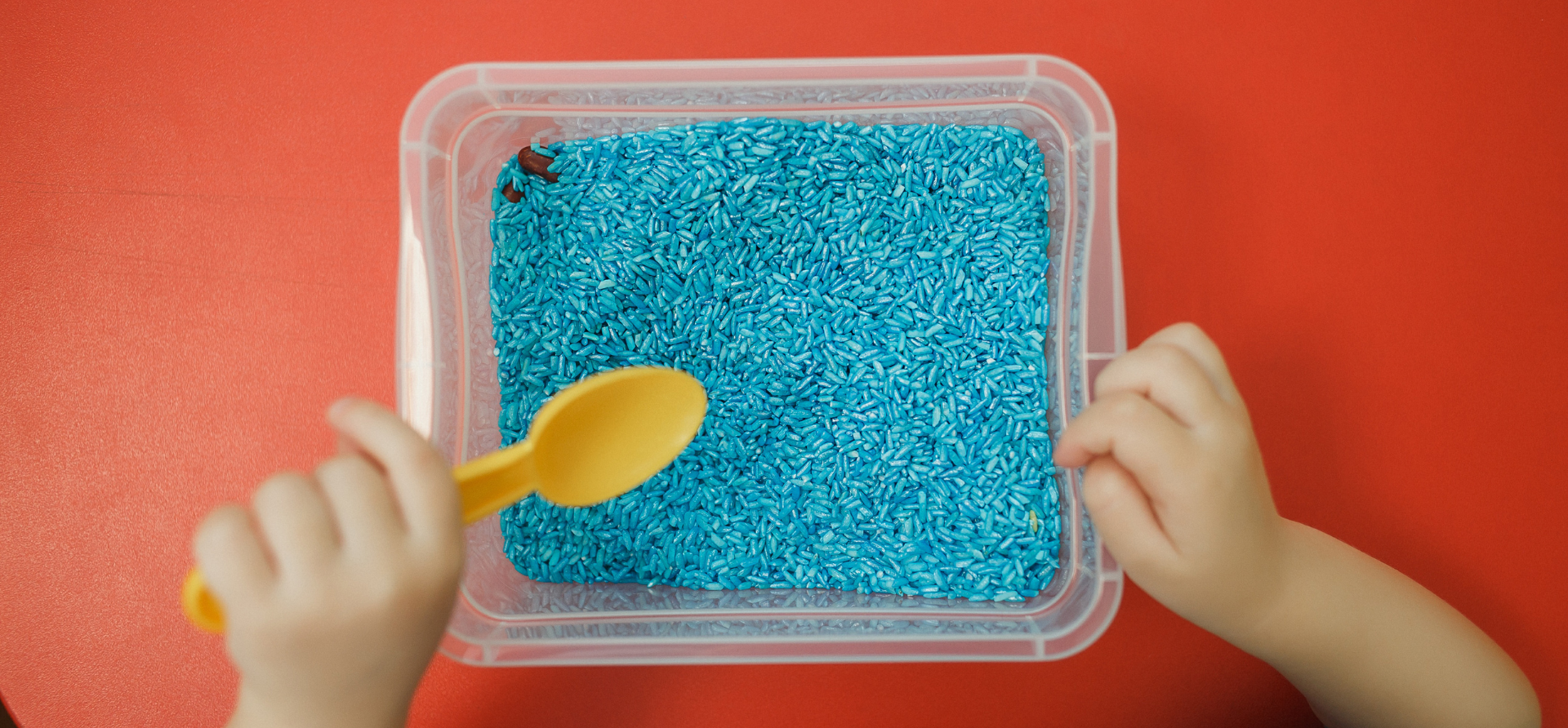 Child's hands playing with blue glitter in a plastic container, with a yellow spoon on a red surface.
