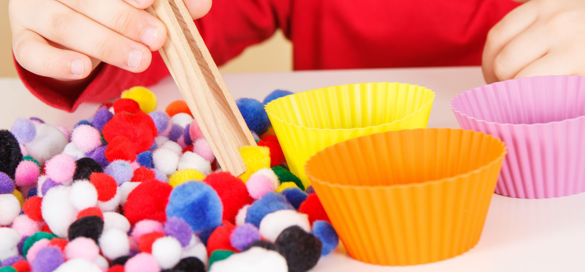 Child using tongs to sort colorful pom-poms into orange, yellow, and pink cupcake liners.