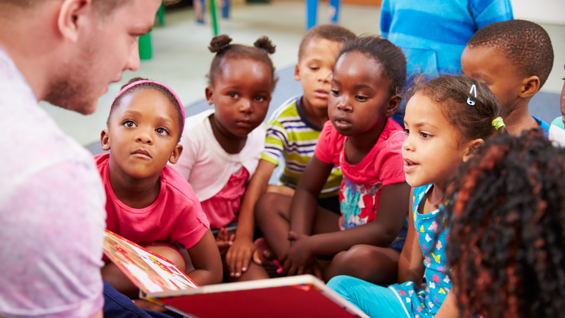 A group of children are sitting on the floor listening to a man read a book.
